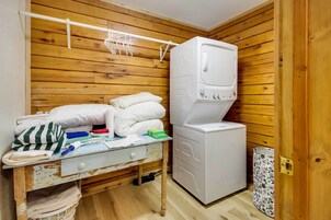 Habitación pequeña con pared de madera, cama con almohadas y ropa doblada, lavadora y una mesita con productos de limpieza.