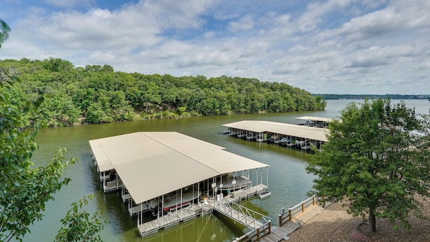 A lakeside property with multiple docked boats, a covered deck area, and a forested shoreline. - Tranquil Osage Beach Condo w/ Waterfront Decks! (Osage Beach)