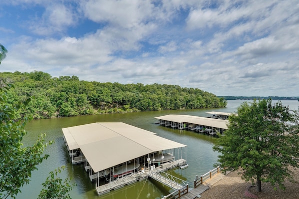 A lakeside property with multiple docked boats, a covered deck area, and a forested shoreline. - Tranquil Osage Beach Condo w/ Waterfront Decks! (Osage Beach)
