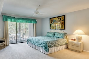 A bedroom with a bed, a nightstand, a lamp, a ceiling fan, a window with curtains, and a balcony with outdoor furniture.