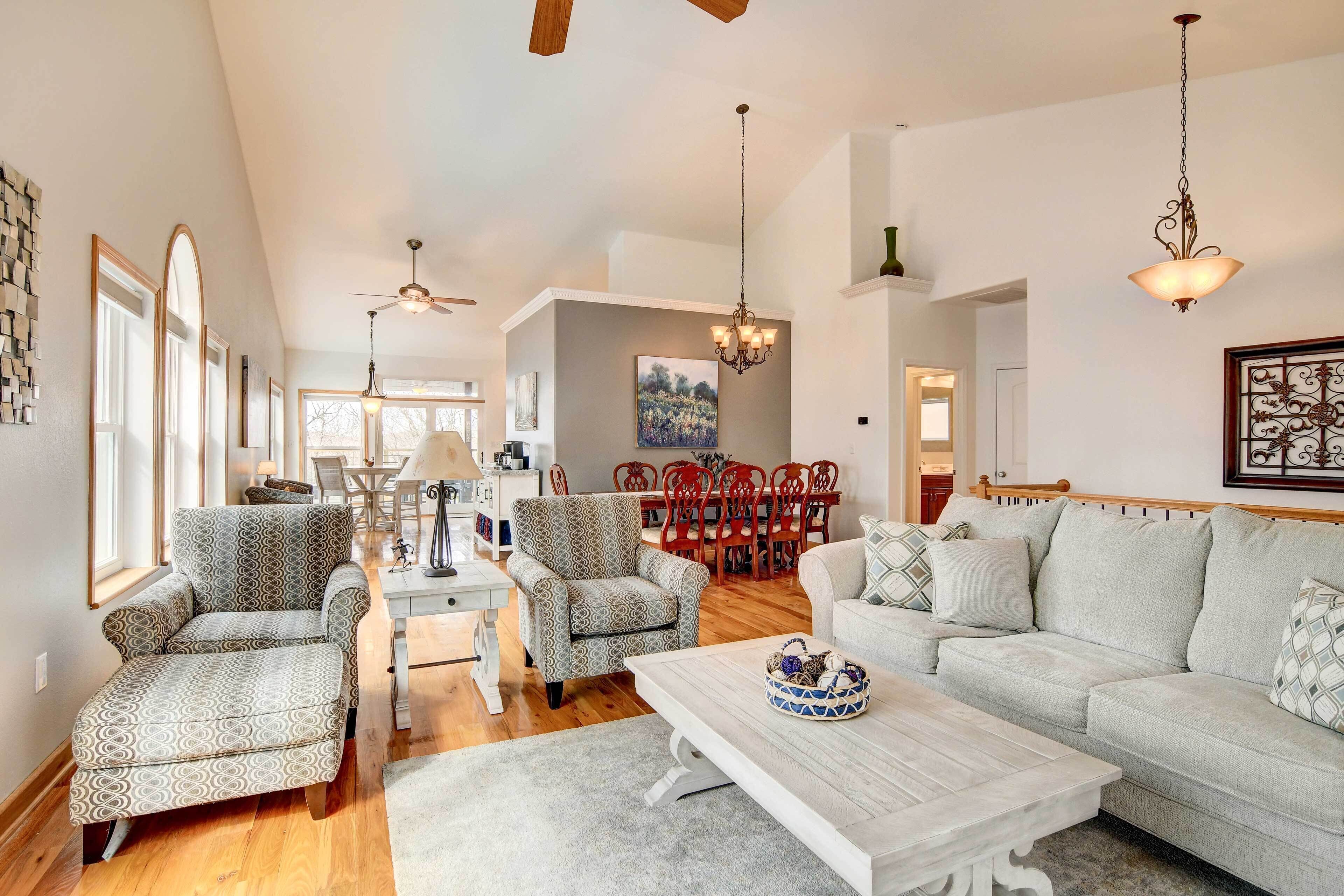 A spacious living room with a large sofa, two patterned armchairs, a coffee table, and a dining area with a red table and chairs.