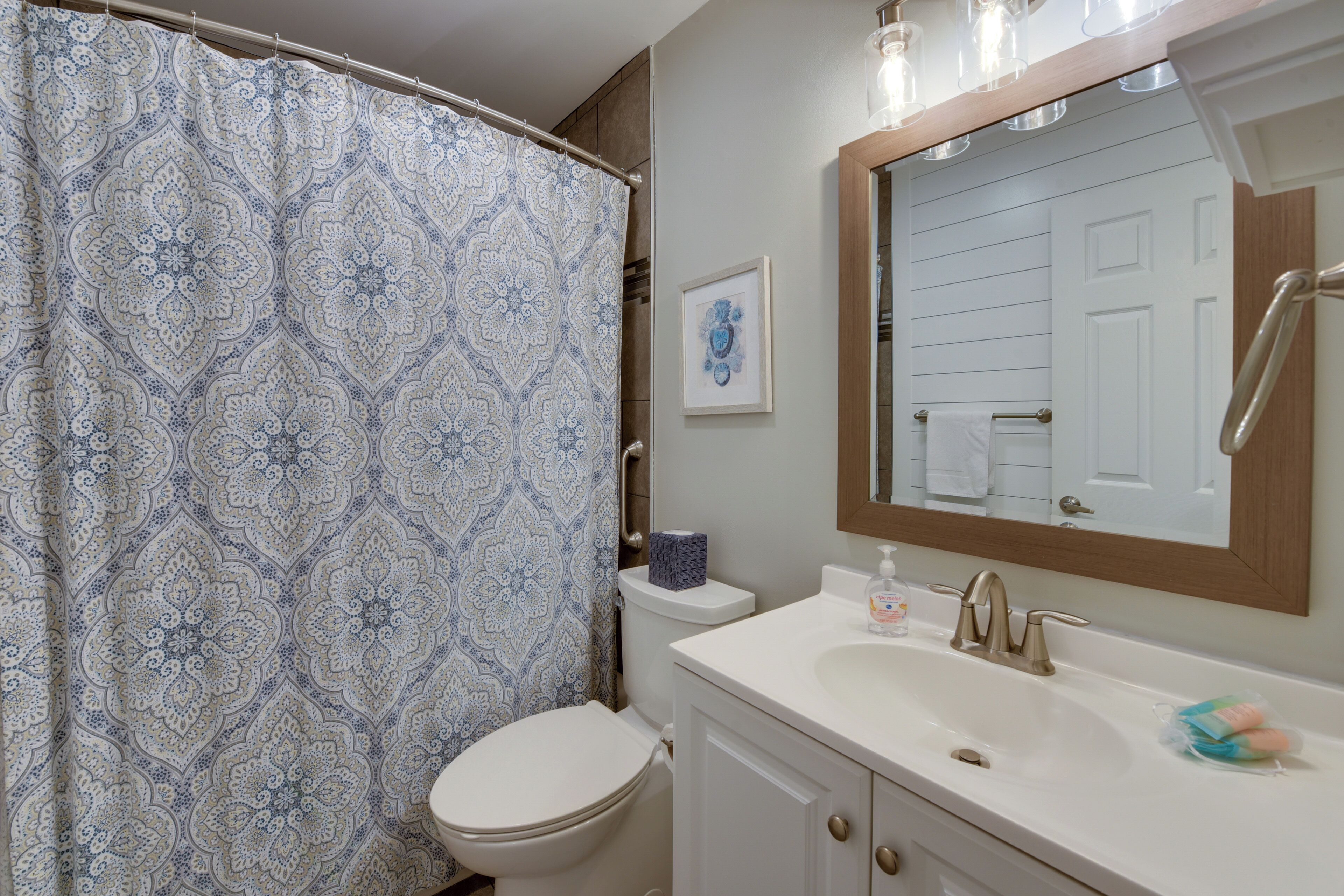 Un baño con cortina de ducha estampada, un inodoro, un lavabo con espejo y un cuadro en la pared.