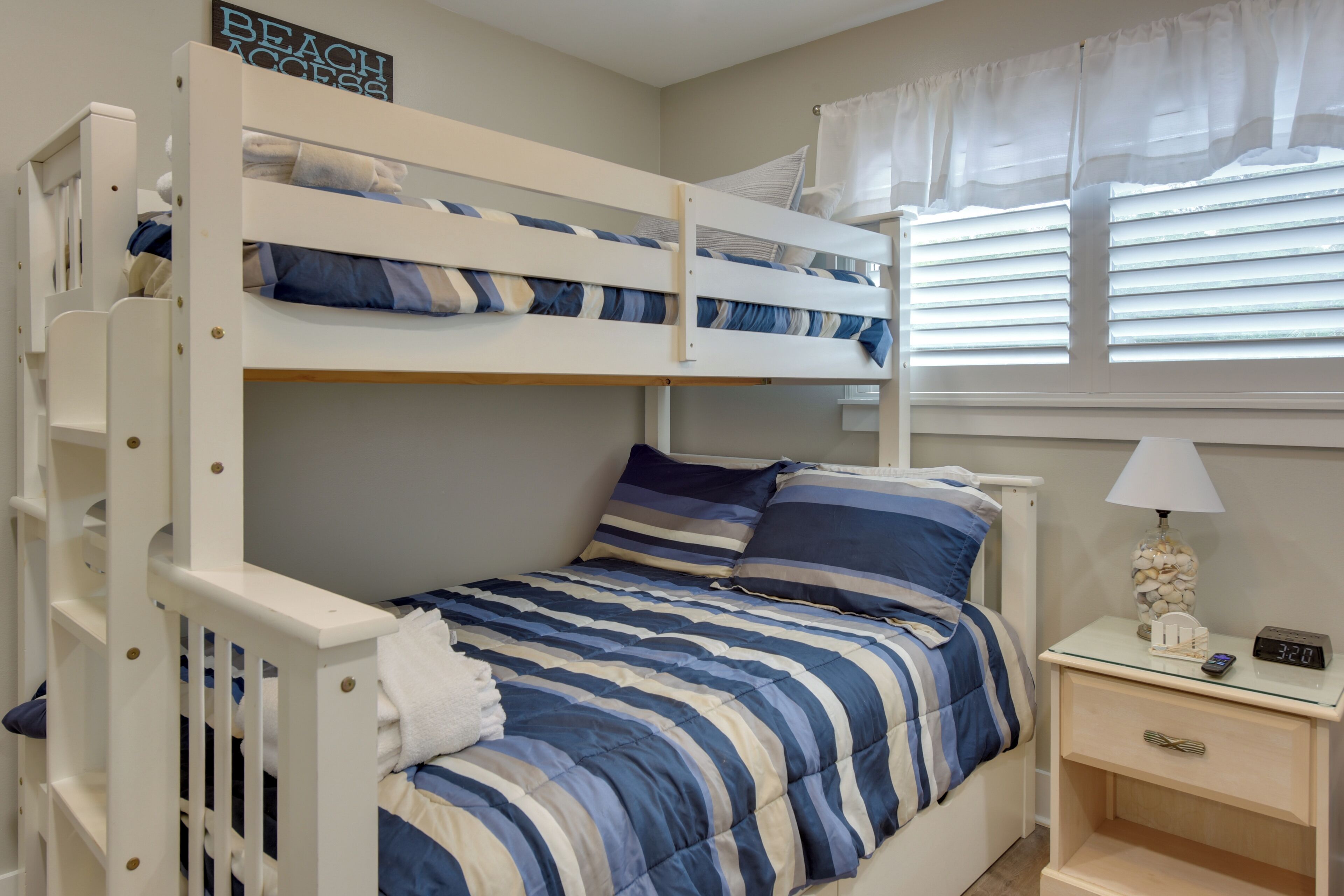 Een stapelbed met blauw-wit gestreept beddengoed, een nachtkastje met een lamp, en een bordje aan de muur met de tekst 'BEACH ACCESS'.