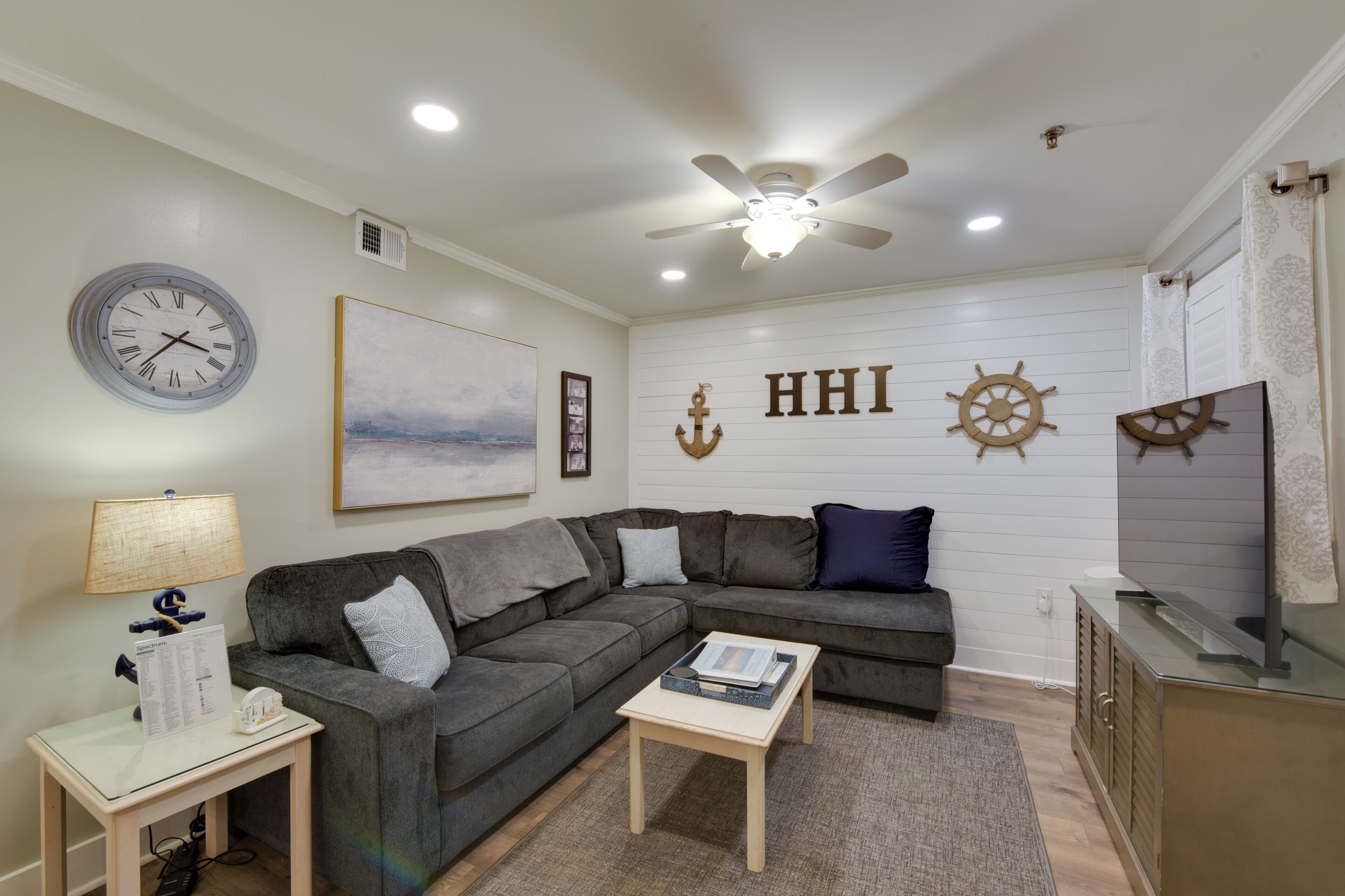 A living room with a sectional sofa, a coffee table, a wall clock, a ceiling fan, and nautical-themed wall decor.