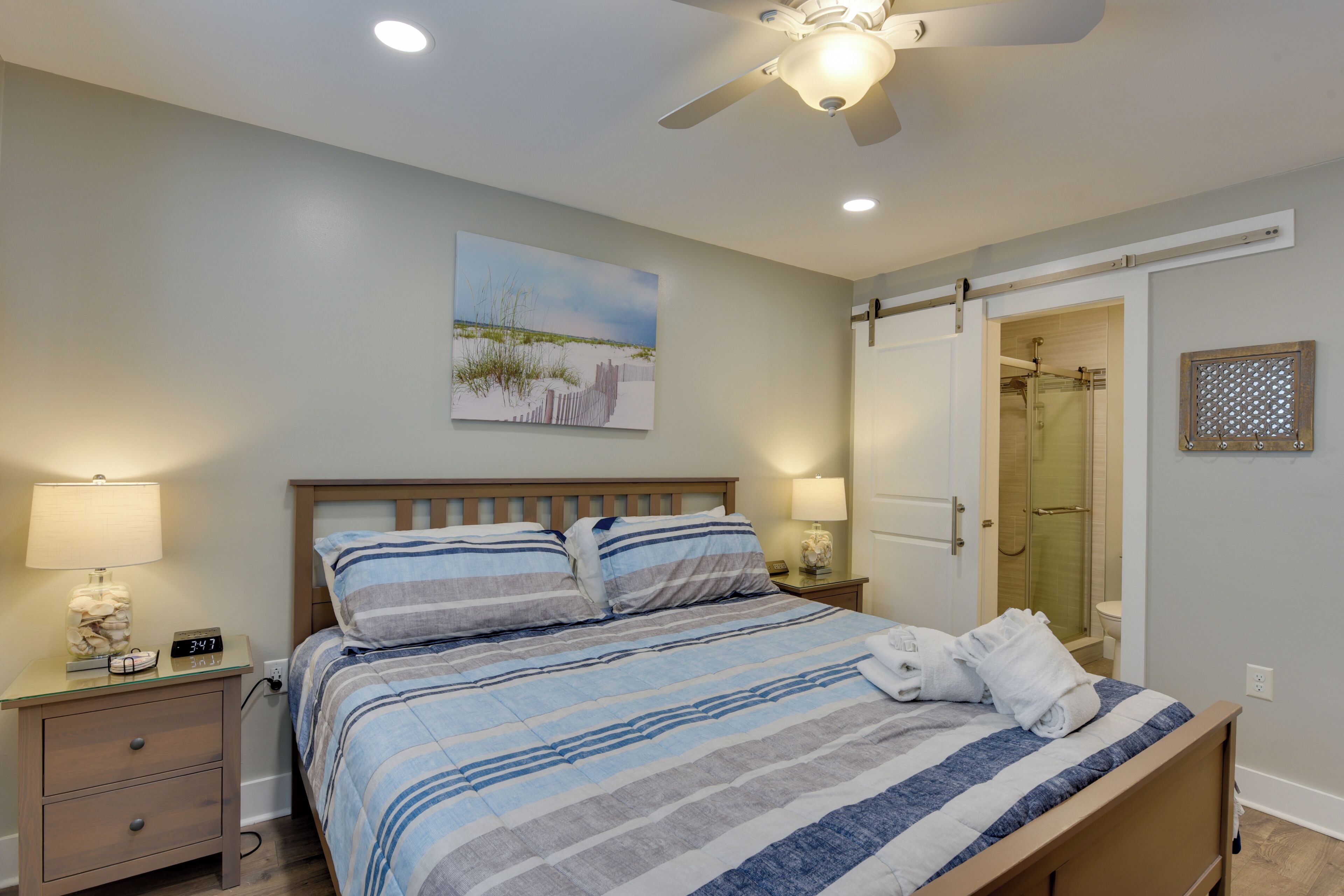 A bedroom with a bed, nightstands, a ceiling fan, a painting, and a door leading to a bathroom.