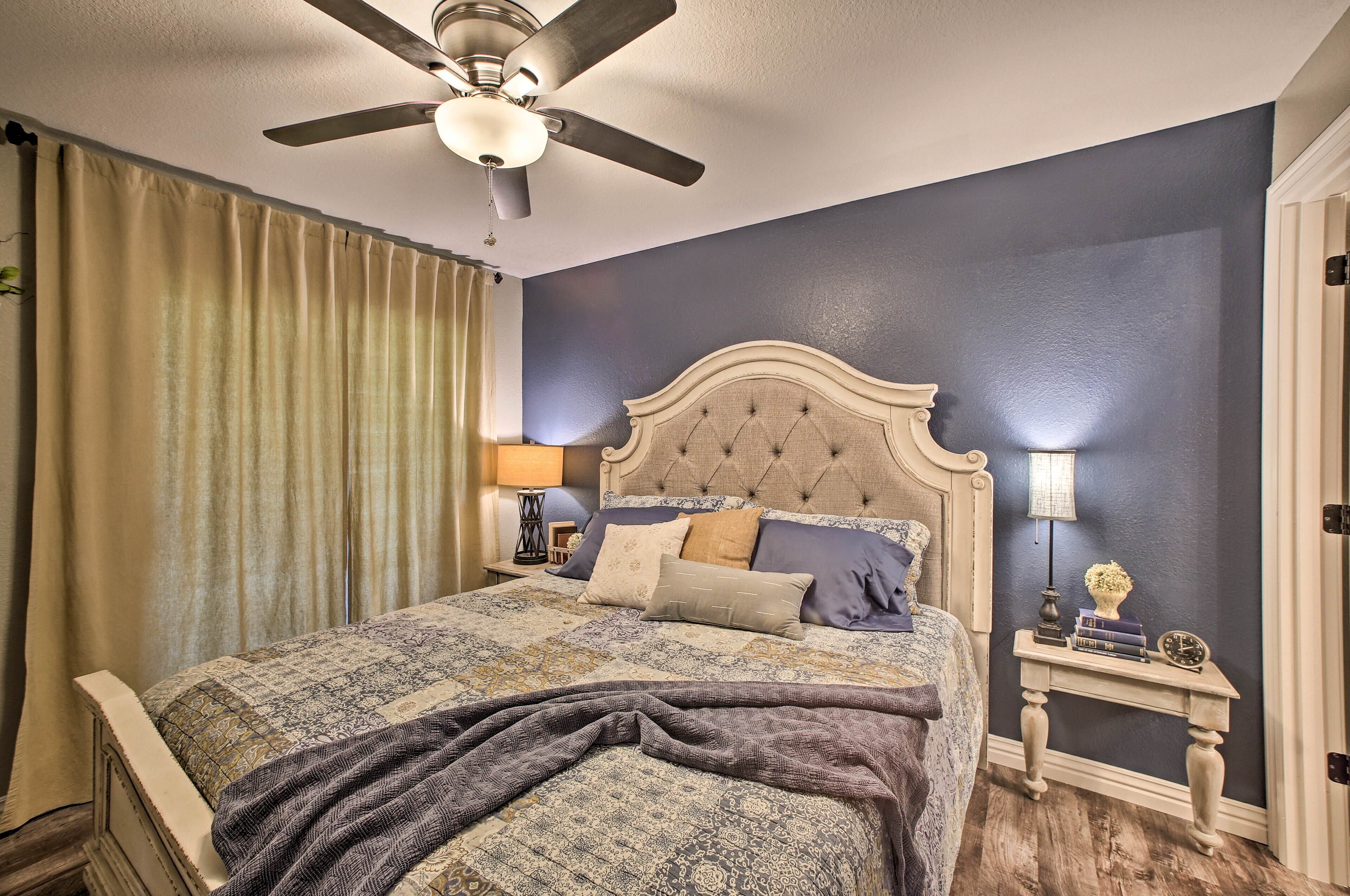 A bedroom with a large bed, a ceiling fan, a nightstand with a lamp, and a door.