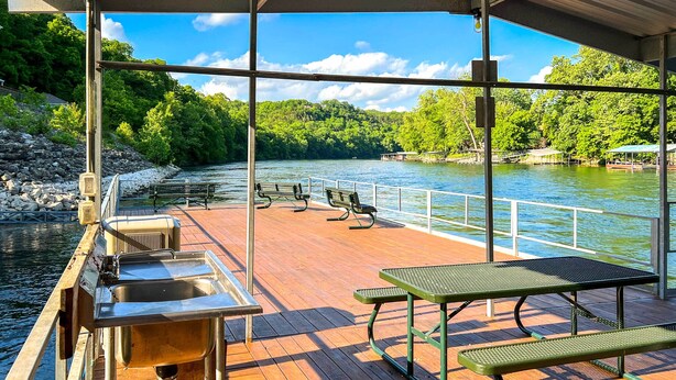 A covered deck with picnic tables and a metal sink, overlooking a river with a forested hillside.