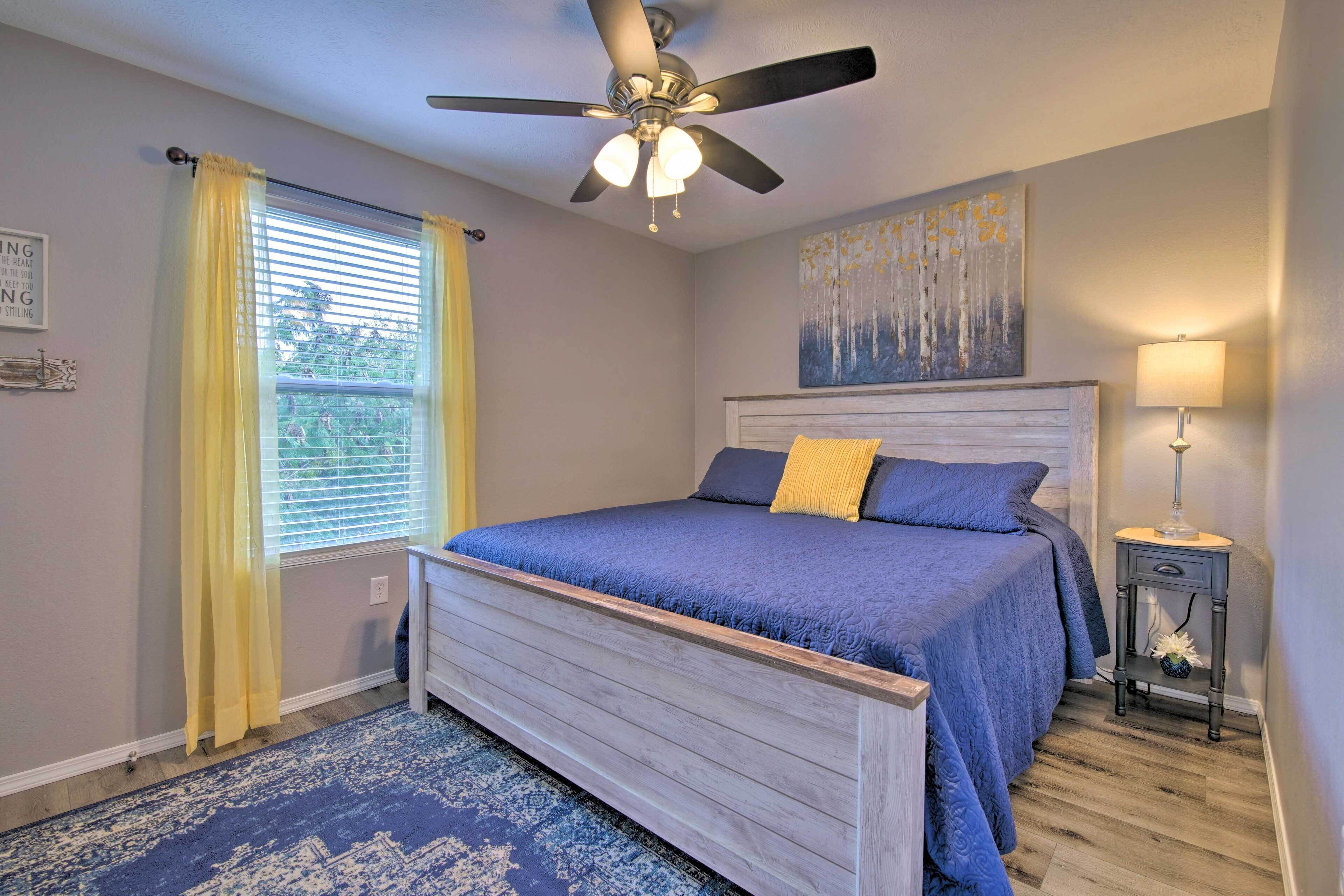 A bedroom with a bed, a ceiling fan, a window with yellow curtains, a bedside table with a lamp, and a wall with a painting.