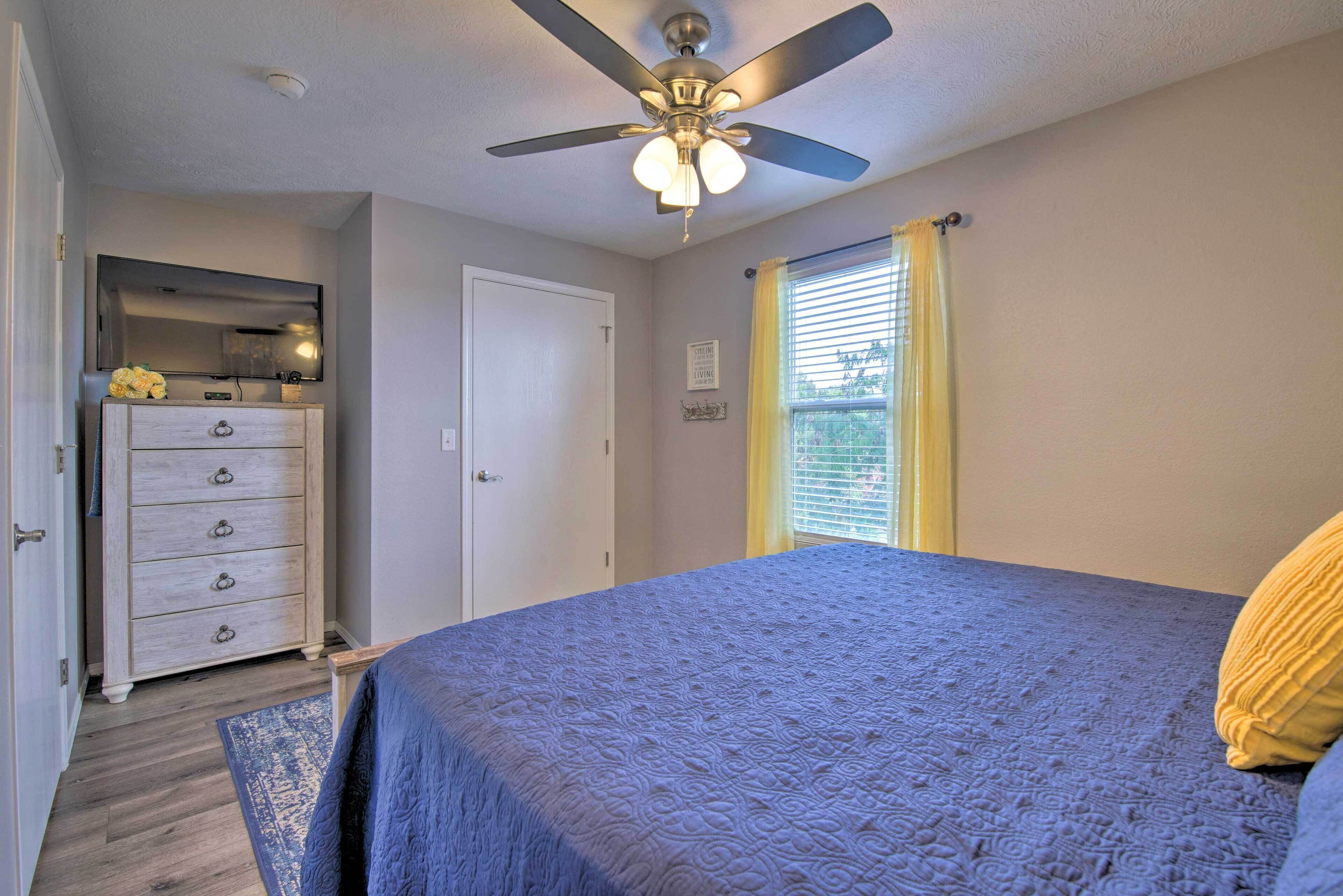 A bedroom with a bed, a ceiling fan, a dresser, a window with curtains, and a door.