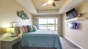 A bedroom with a bed, a nightstand, a ceiling fan, a TV on a shelf, and a view of the outdoors. - Community Perks! Waterfront Lake Ozark Escape (Lake Ozark)