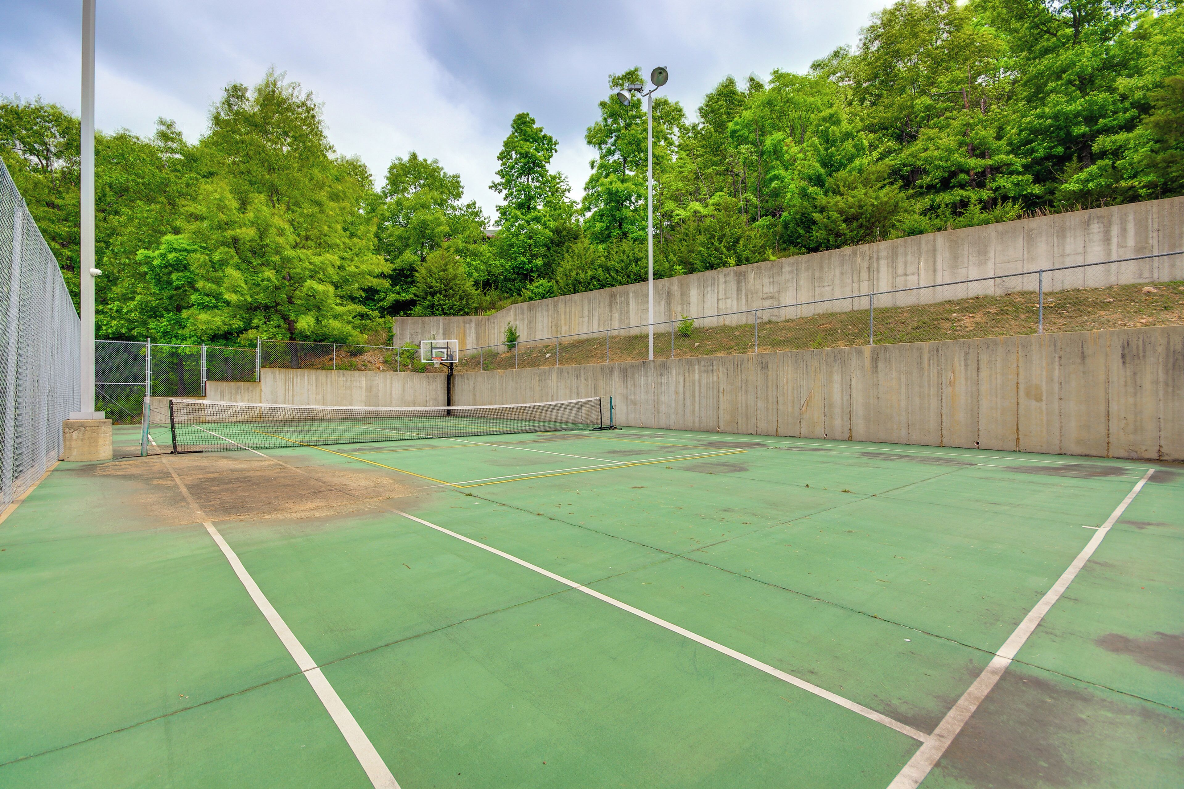 Een binnentennisbaan met een net, omgeven door een betonnen muur en bomen.