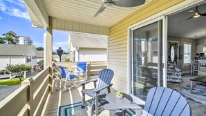 A covered outdoor deck with a ceiling fan, two Adirondack chairs, a small table, and a glass door leading to an interior space. - Updated Myrtle Beach Cottage w/ Shared Pool! (Myrtle Beach)