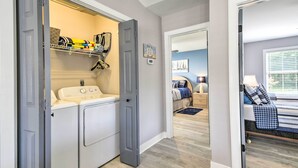 A laundry room with a washing machine and dryer, a shelf with folded clothes, and a door leading to a bedroom with a bed and a nightstand. - Updated Myrtle Beach Cottage w/ Shared Pool! (Myrtle Beach)