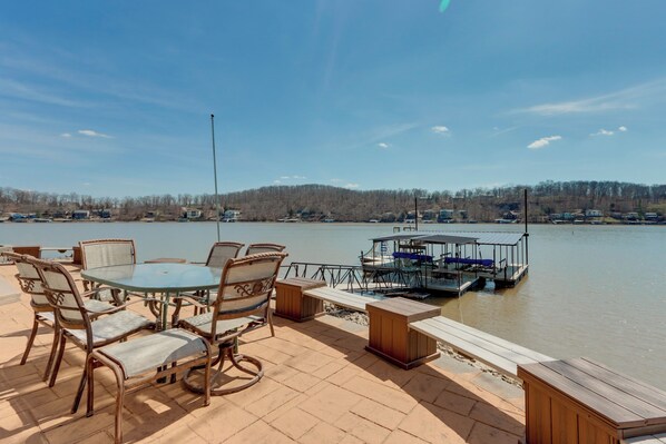 A patio with a round table and chairs, a dock with a boat, and a lake view.