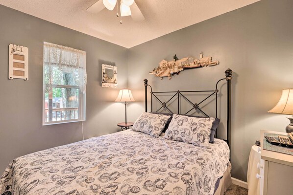A bedroom with a bed, bedside lamps, a nightstand, a window with lace curtains, and a wall-mounted shelf with decorative items. - Walk to Waterfront: Osage Beach Escape! (Osage Beach)