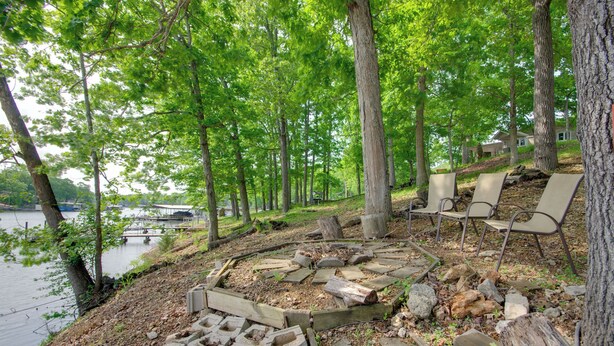 A lakeside deck with stone seating and a fire pit, surrounded by trees and a view of a house across the water. - Waterfront Lake of the Ozarks Home w/ Views! (Gravois Mills)