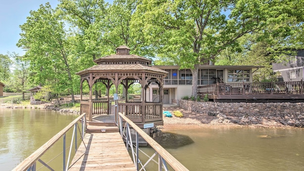 A wooden pier leading to a gazebo by a calm river, with a house and dock in the background.