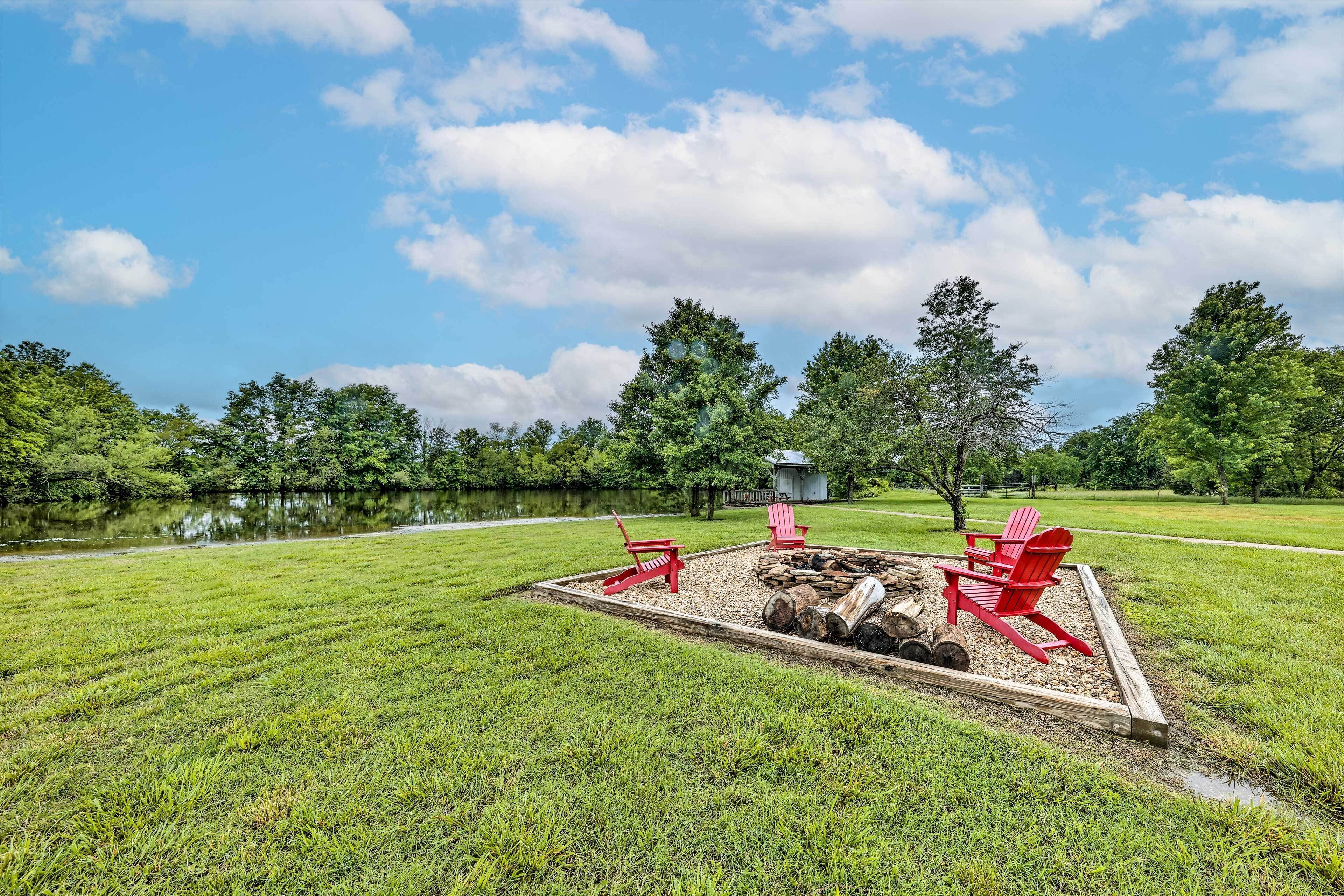 A fire pit area with red Adirondack chairs, surrounded by a wooden deck, set in a grassy field with a lake and trees in the background.