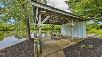 A rustic wooden pavilion with a picnic table, situated by a calm lake, surrounded by lush greenery.
