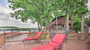 A wooden deck with two lounge chairs and a table, overlooking a body of water and a cabin. - Hot Tub, Dock! Waterfront Lake Ozark Home! (Lake Ozark)