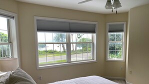 A bedroom with a large bed, two windows with blinds, a ceiling fan, and a view of a road and greenery outside.