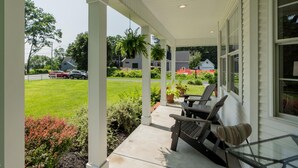 A covered porch with two chairs and a table, overlooking a green lawn and a street with parked cars.