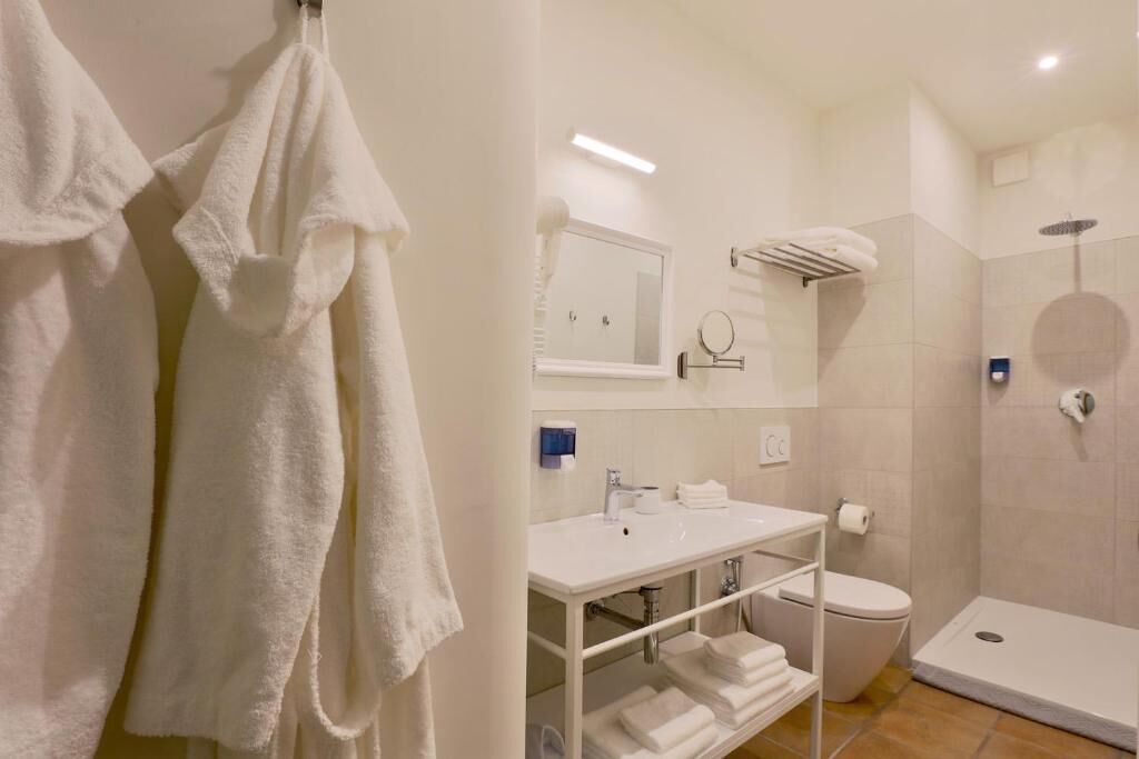A modern bathroom with a white sink, toilet, and shower. There are towels hanging on the wall and a shelf with folded towels.