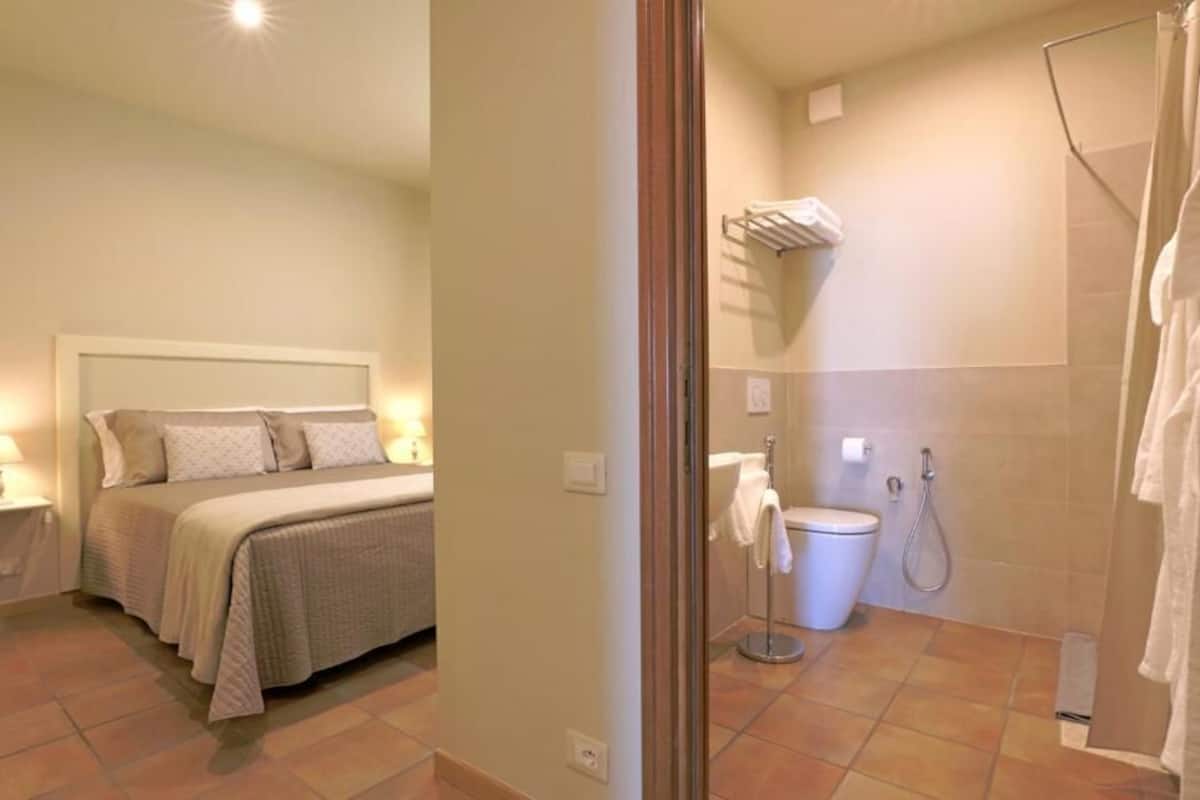A bedroom with a bed, bedside table, and a bathroom with a shower, toilet, and towel rack.