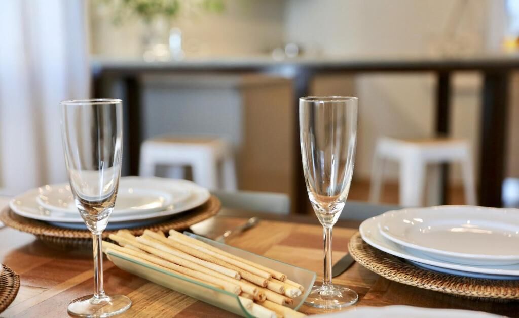 Una mesa de comedor dispuesta con dos copas de vino, platos y un bol de palitos de vidrio.