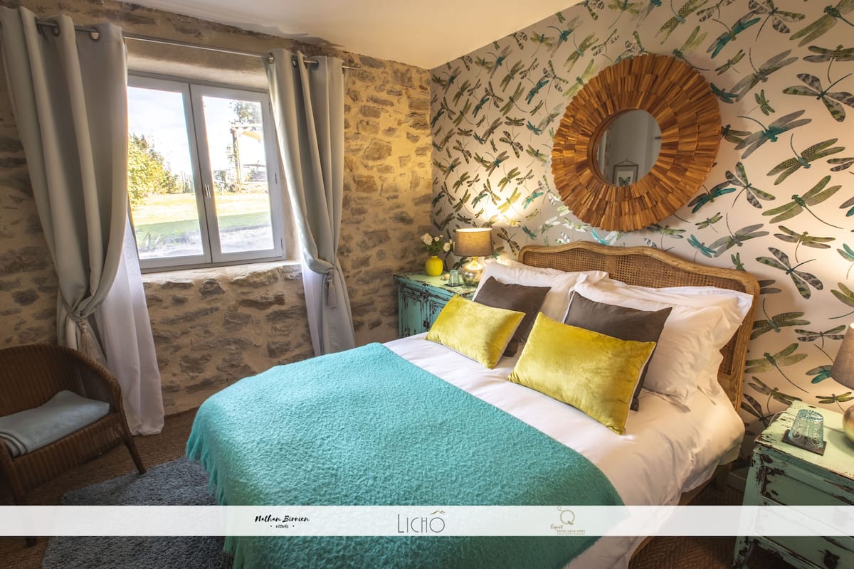 Une chambre avec un mur en pierre, un lit avec une literie aux couleurs turquoise et jaune, une chaise en osier, une table de chevet avec une lampe et une fenêtre avec des rideaux.