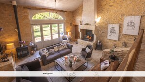 A spacious living room with a stone fireplace, comfortable seating, and a large window offering a view of the outdoors. - Maison des Quatre Saisons (Les Esseintes)