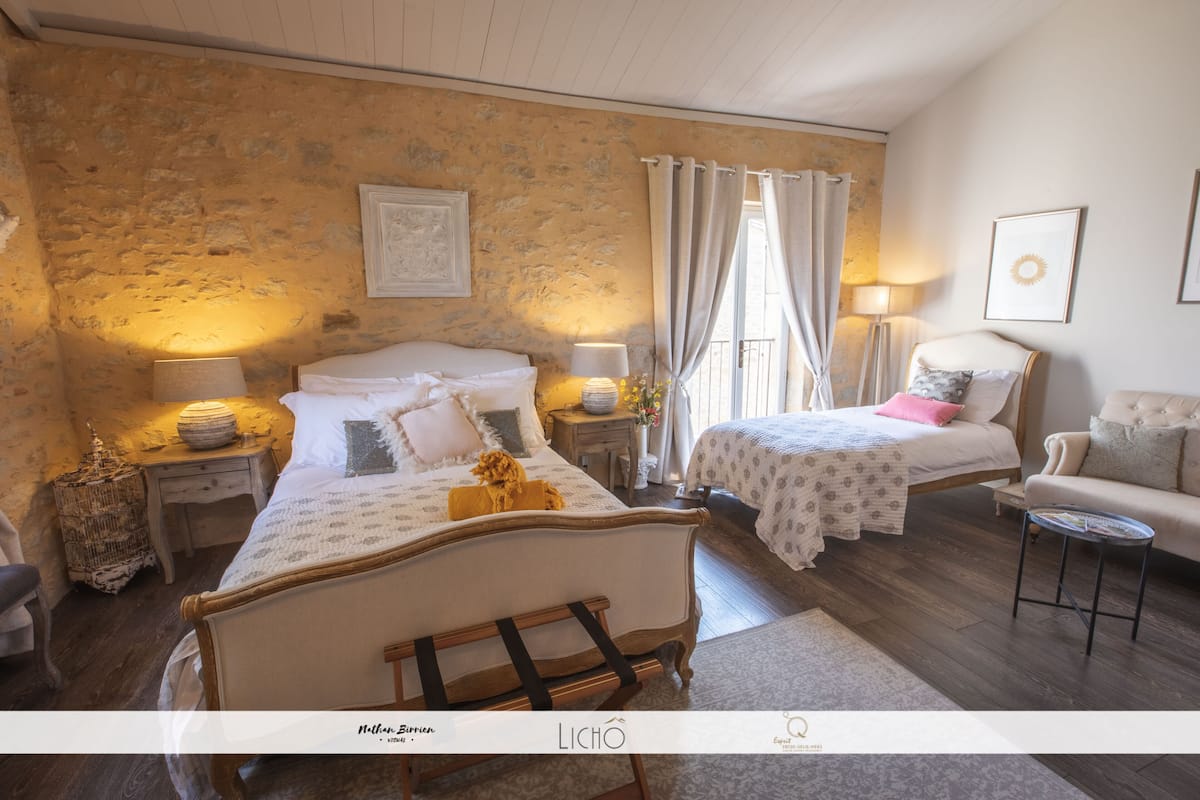 Une chambre avec un mur en pierre, deux lits, une chaise, une petite table et une lampe.