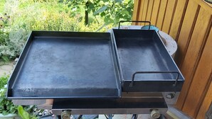 A barbecue grill with a flat cooking surface and a side tray, set on a wooden deck with a garden in the background.