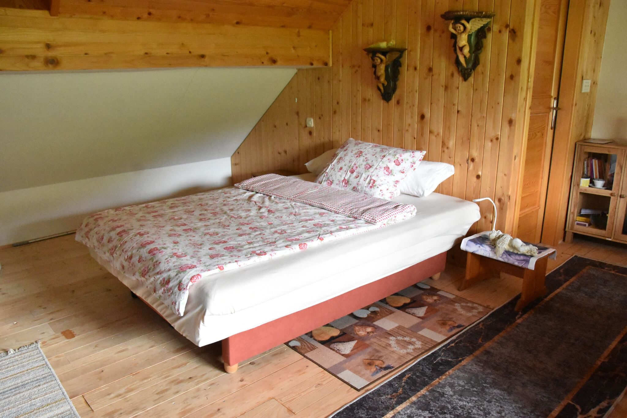 A bedroom with a wooden ceiling, a bed with floral bedding, a small bedside table, and a bookshelf.