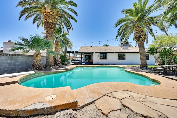 A swimming pool with a stone deck, surrounded by palm trees and a house with a covered patio.