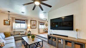 A modern living room with a sectional sofa, a flat-screen TV mounted on the wall, a ceiling fan, and a large window with blinds. - Yuma Vacation Rental w/ Private Pool & Patio! (Yuma)