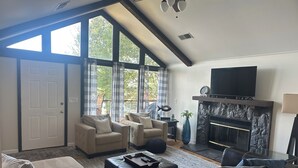 Ein geräumiges Wohnzimmer mit Kamin, großen Fenstern und Deckenventilator.