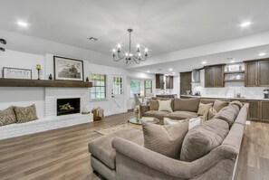 A spacious living room with a fireplace, a large beige sectional sofa, and a kitchen in the background. - Luxurious & COZY South Tulsa Stunning ReDesign (Tulsa)