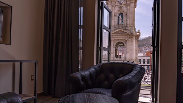 Ein Zimmer mit Ledersessel und kleinem runden Tisch, Blick auf ein historisches Gebäude durch eine offene Tür sowie ein Balkongeländer.