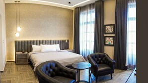 Ein Hotelzimmer mit einem großen Bett, zwei Nachttischen, einer modernen Kronleuchter, einem kleinen runden Tisch, zwei Ledersesseln und einem großen Fenster mit Vorhängen.