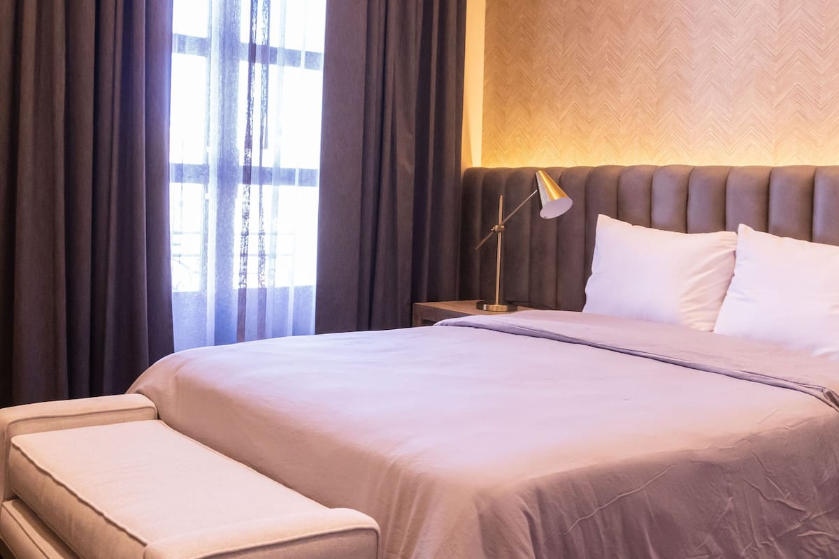 Een hotelkamer met een groot bed, een nachtkastje met een lamp, een klein bankje en een bedtafeltje met een telefoon.