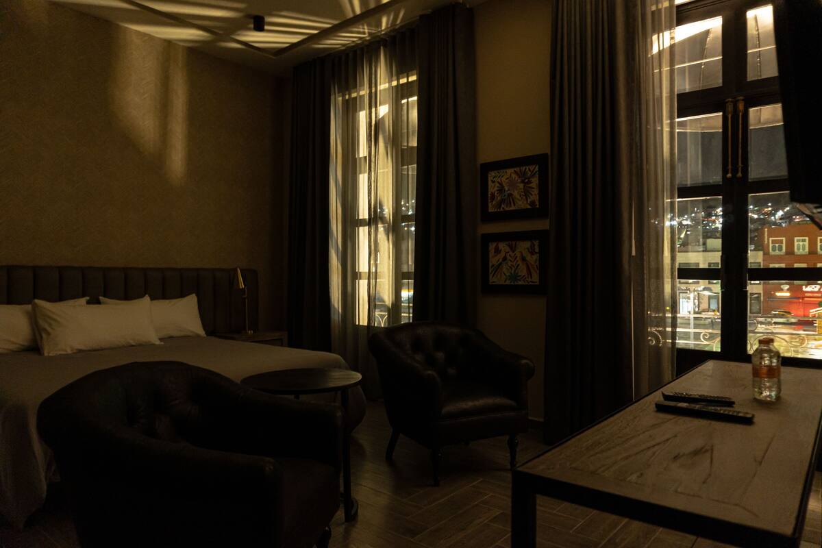 Een hotelkamer met een bed, een stoel, een klein tafeltje en uitzicht op de stad door grote ramen.
