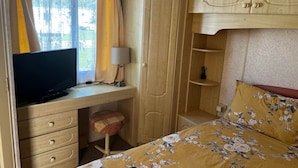 A bedroom with a bed, a desk with a TV, a lamp, and wooden cabinets. - 2 Bedroom Caravan Hunstanton Pets go Free (Hunstanton)