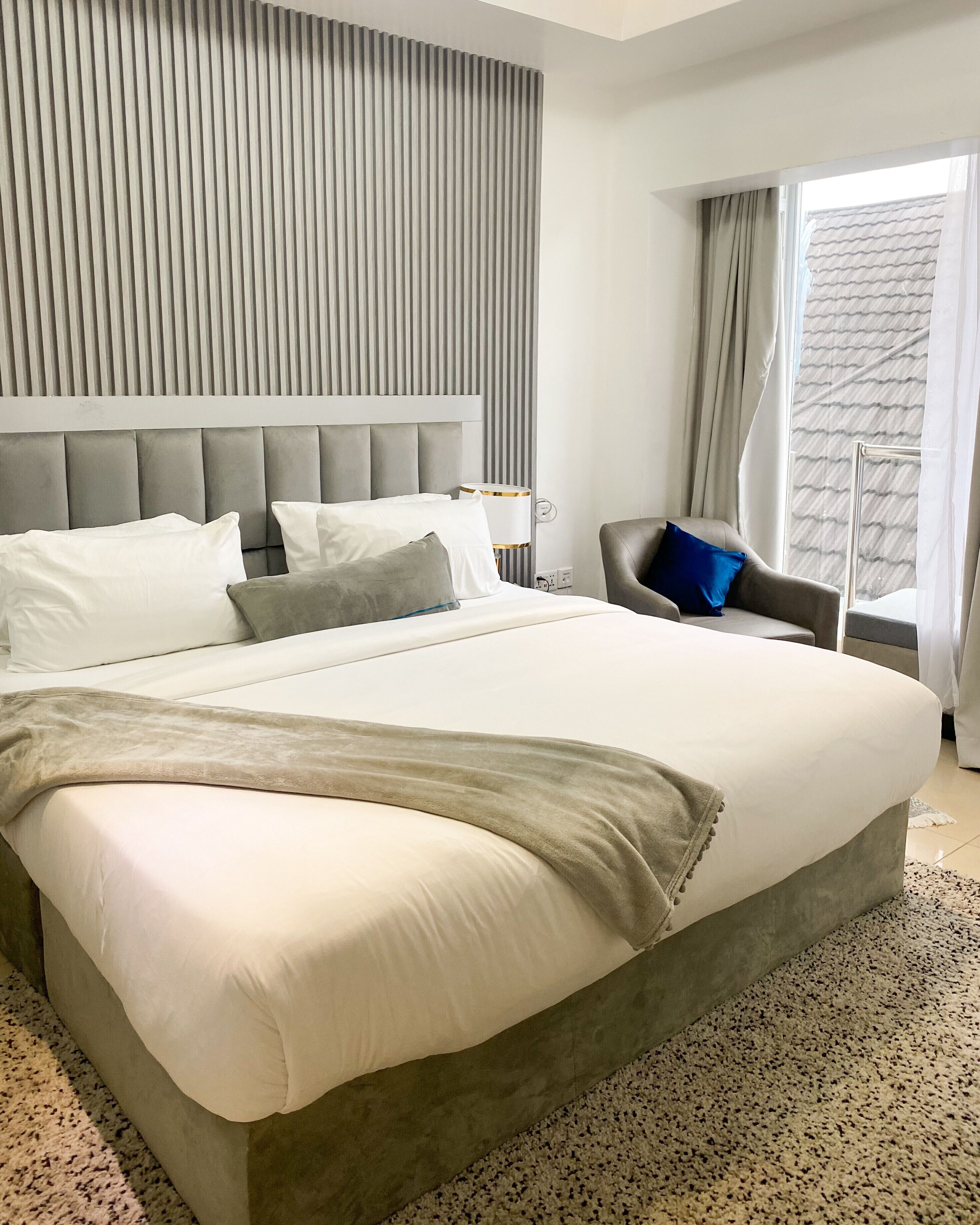 Una habitación de hotel moderna con una cama grande, un cabecero tapizado en gris, una silla con un cojín azul y una ventana con cortinas.
