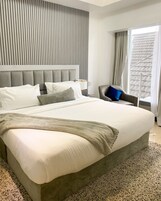 Una habitación de hotel moderna con una cama grande, un cabecero tapizado en gris, una silla con un cojín azul y una ventana con cortinas.