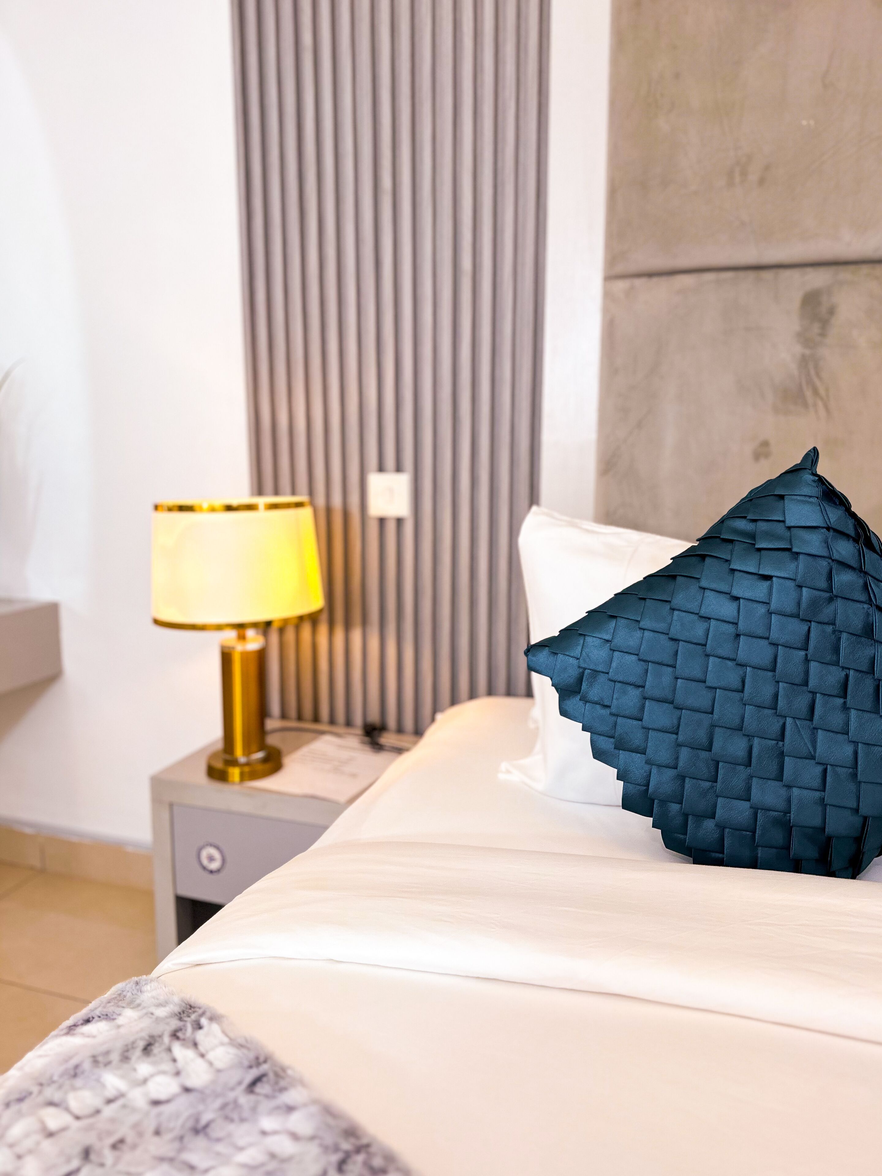 A neatly made bed with a white comforter and a dark blue woven pillow. A gold-colored lamp is on a bedside table next to the bed.