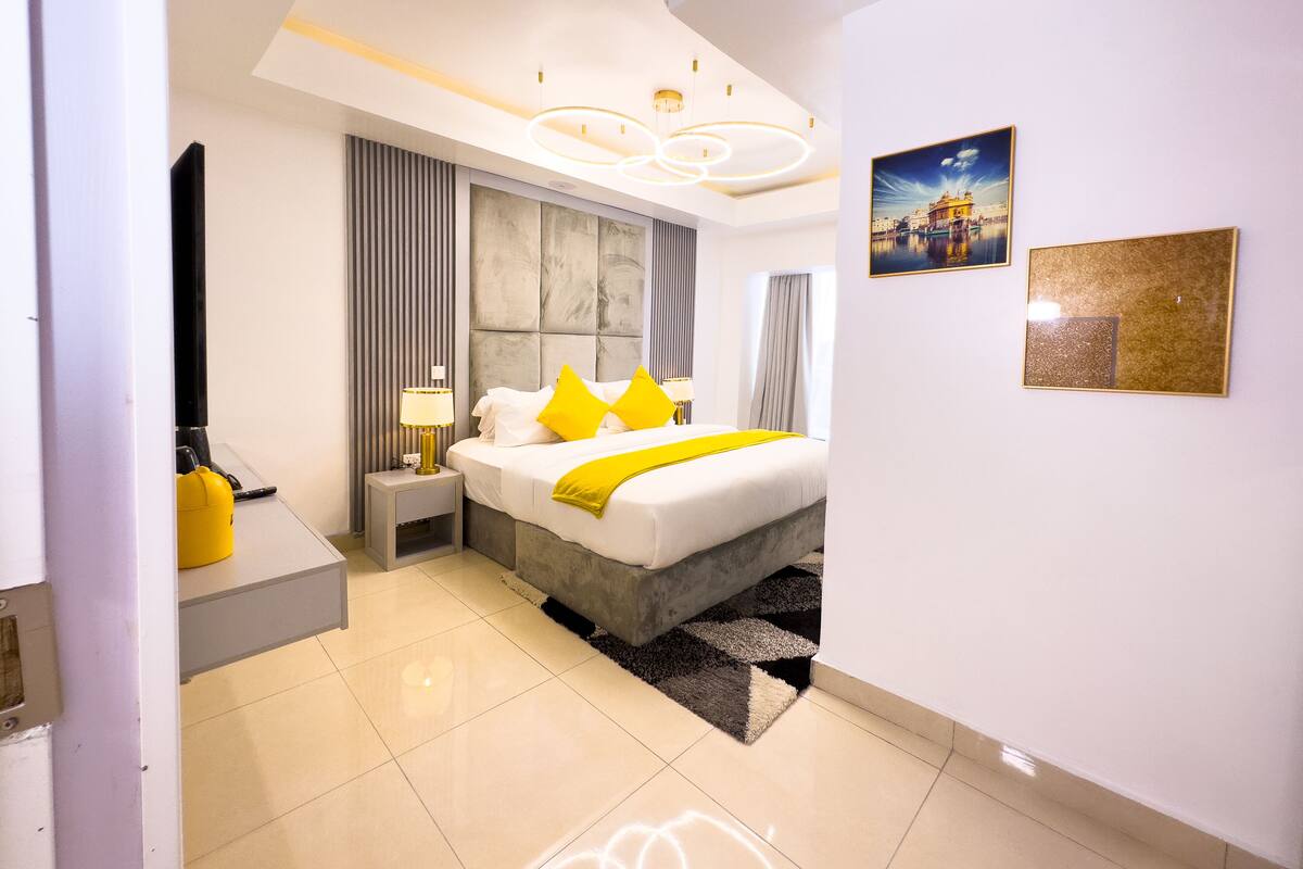 A modern hotel room with a large bed, a nightstand, a lamp, a picture on the wall, and a view of a building outside.