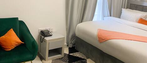A modern hotel room with a bed, a green armchair, a small bedside table, and a wall-mounted phone.