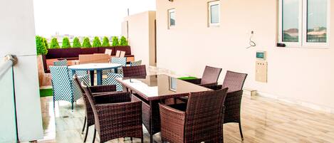 A modern outdoor dining area with wicker furniture, a glass table, and a view of greenery and buildings in the background.