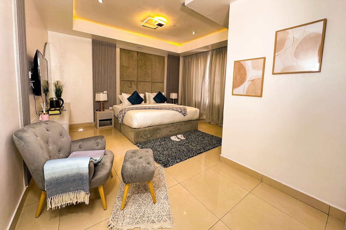 A modern hotel room with a large bed, a grey armchair, a small ottoman, a flat-screen TV, and a bedside table with a lamp and a decorative item.